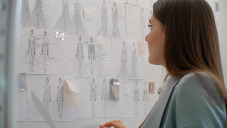 Young-fashion-designer-standing-with-back-to-camera-looking-at-drawings-sketches-hanging-on-wall.-Woman-is-thinking-about-new-trendy-clothing-collection